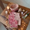 Cotons démaquillants lavables Noël rouges et beiges avec panière sur table en bois Cotons démaquillants lavables Noël rouges à motif nordique et beiges à flocons avec leur panière, posés sur une table en bois avec bougies, flacons de soin et guirlande lumineuse, cousus main par Atelier Julac