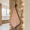 Essuie-mains éponge coton imprimé Noël motif flocons suspendu Essuie-mains éponge Atelier Julac en coton imprimé Noël, motif flocons rouges sur fond écru, suspendu à un crochet dans une salle de bain en bois avec guirlande lumineuse