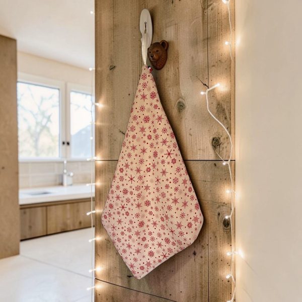 Essuie-mains éponge coton imprimé Noël motif flocons suspendu Essuie-mains éponge Atelier Julac en coton imprimé Noël, motif flocons rouges sur fond écru, suspendu à un crochet dans une salle de bain en bois avec guirlande lumineuse
