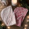 Serviettes cheveux bambou Noël rouge et beige chalet montagne Serviette cheveux bambou Noël, une beige à flocons rouges et une rouge motif nordique, posées avec bougie, branches de sapin et guirlande lumineuse, cousues main par Atelier Julac