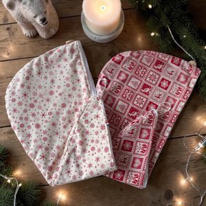 Serviette cheveux bambou Noël, une beige à flocons rouges et une rouge motif nordique, posées avec bougie, branches de sapin et guirlande lumineuse, cousues main par Atelier Julac