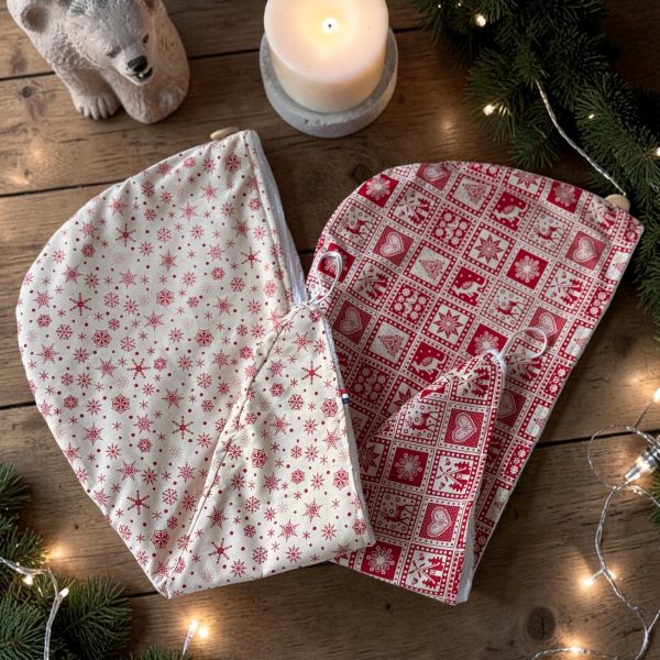 Serviettes cheveux bambou Noël rouge et beige chalet montagne Serviette cheveux bambou Noël, une beige à flocons rouges et une rouge motif nordique, posées avec bougie, branches de sapin et guirlande lumineuse, cousues main par Atelier Julac
